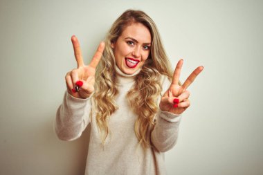 İzole edilmiş beyaz arka planda boğazlı kazak giyen güzel bir kadın her iki elin parmaklarını zafer işareti yaparken göstererek gülümsüyor. İki numara..