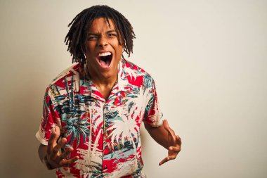 Tatilde rastalı Afro adam izole edilmiş beyaz arka plan üzerine yazlık tişört giyiyor çılgın ve sinirli bir şekilde bağırıyor ve agresif bir ifade ve kollar havada bağırıyor. Hayal kırıklığı konsepti.