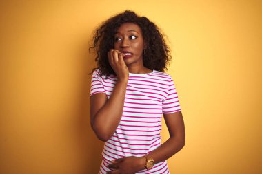 İzole edilmiş sarı arka plan üzerinde çizgili tişört giyen genç Afro-Amerikalı kadın elleri ağız yiyen tırnaklarla stresli ve gergin görünüyor. Endişe sorunu.
