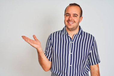 Mavi çizgili gömlekli genç adam izole edilmiş beyaz arka planda dikiliyor neşeli bir şekilde gülümsüyor ve avuç içiyle kameraya bakıyor..