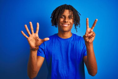 Rastalı tişörtlü Afro-Amerikalı adam izole edilmiş mavi arka plan üzerinde duruyor ve yedinci parmaklarını gösterirken kendinden emin ve mutlu bir şekilde gülümsüyor..