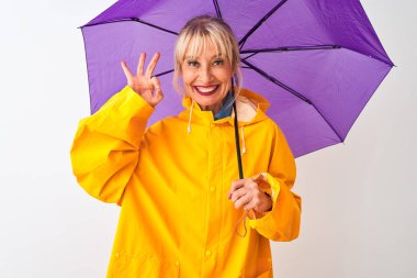 İzole edilmiş beyaz arkaplanda orta yaşlı kadın yağmurluk ve mor şemsiye giyiyor. Tamam işareti yapıyor, mükemmel bir sembol.