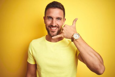 Sarı izole edilmiş arka plan üzerine gündelik sarı tişört giyen genç yakışıklı adam telefonla el ve parmaklarıyla telefonla konuşuyor gibi gülümsüyor. İletişim kavramları.
