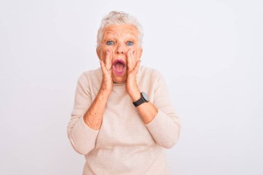 Yaşlı, gri saçlı, balıkçı yaka kazak giyen bir kadın izole edilmiş beyaz arka planda korku ve şaşkınlık içinde duruyor.