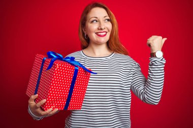 Kızıl saçlı güzel bir kadın kırmızı izole edilmiş arka planda doğum günü hediyesini tutuyor ve gülümseyen bir gülümsemeyle baş parmağıyla işaret ediyor.