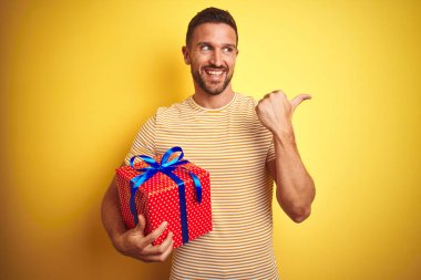 Sarı arka planda bir doğum günü hediyesi tutan genç adam başparmağıyla gülümseyerek yüzünü gösteriyor.