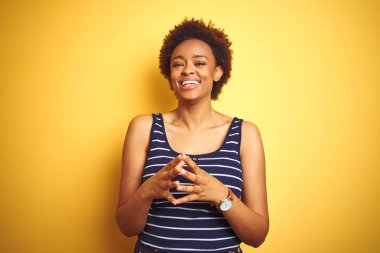 Beauitul Afro-Amerikalı kadın izole edilmiş sarı arka plan üzerine yaz tişörtü giyiyor ellerini birleştirip gülümseyerek gevşemiş ve neşeli gülümsüyor. Başarı ve iyimser