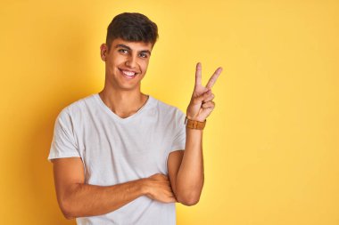 İzole edilmiş sarı arka planda duran beyaz tişörtlü genç Hintli adam kameraya gülümseyen ve parmaklarıyla zafer işareti yapan. İki numara..