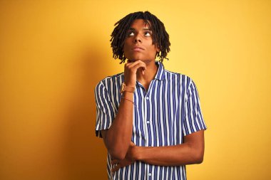 Rastgele çizgili tişört giyen Afro bir adam izole edilmiş sarı arka plan üzerinde elleri çenesinde soru sormayı düşünen düşünceli bir yüz ifadesi var. Düşünceli bir yüzle gülümsüyor. Şüphe kavramı.