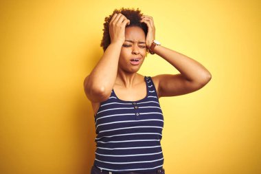 Beauitul Afro-Amerikalı kadın izole edilmiş sarı arka plan üzerine yaz tişörtü giyiyor. Ağrı ve migrenden dolayı çaresiz ve stresli. Ellerini başının üstüne koy..