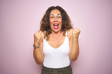 Pembe izole edilmiş arka planda duran orta yaşlı bir kadın kollarını kaldırıp gözleri açık bir şekilde başarıyı kutluyor ve şaşırıyor. Kazanan konsept.