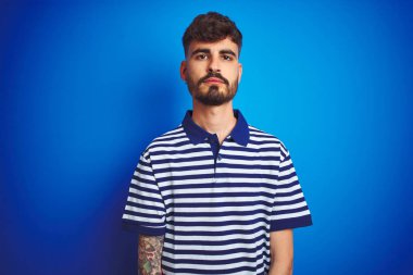 İzole edilmiş mavi arka planda duran çizgili polo giyen genç adam yüzünde ciddi bir ifadeyle rahatlamış. Basit ve doğal bir şekilde kameraya bakıyorsun..