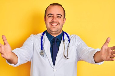 Paltolu ve steteskoplu genç bir doktor izole edilmiş sarı arka planda durup kucaklaşmak için kollarını açıp gülümseyen kameraya bakıyor. Mutluluğu kucaklayan neşeli bir ifade..