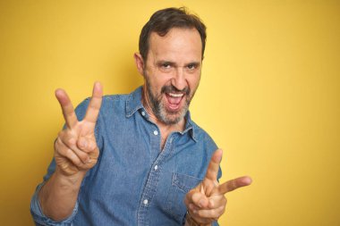 Yakışıklı, orta yaşlı, gri saçlı, izole edilmiş sarı arka planda gülümseyen ve parmaklarını zafer işareti yaparken gösteren bir adam. İki numara..