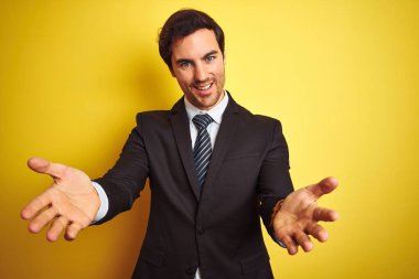 Takım elbiseli ve kravatlı genç yakışıklı işadamı izole edilmiş sarı arka planda dikiliyor neşeli gülümseyerek yardım ve kabullenme eli uzatıyor..