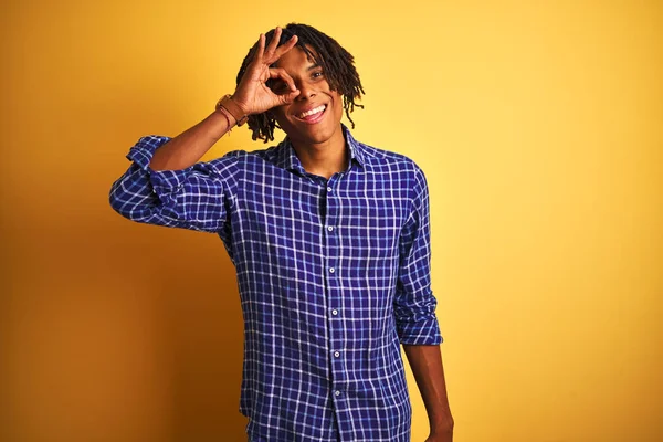 Rastgele gömlek giyen Afro bir adam izole edilmiş sarı arka planda duruyor. El hareketleriyle gülümsüyor, gözleriyle mutlu yüzle parmaklarının arasından bakıyor..