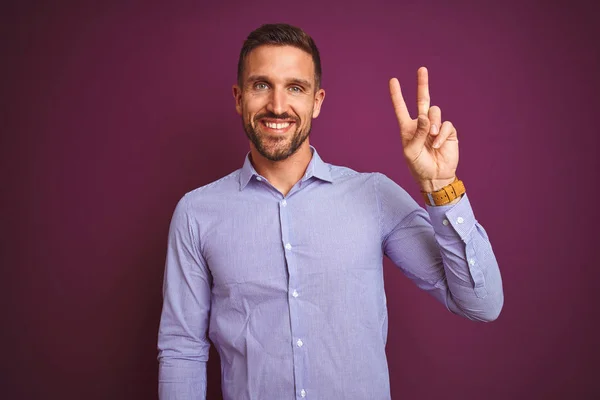 紫色の孤立した背景にエレガントなシャツを着た若いビジネスマンが 勝利のサインをしてカメラに顔をウィンクして幸せな笑顔で笑っています 第二番 — ストック写真