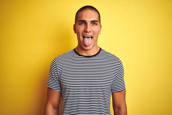 黄色の隔離された背景にストライプのTシャツを着た若いハンサムな男が面白い表情で幸せ舌を突き出しています 感情概念 — ストック写真