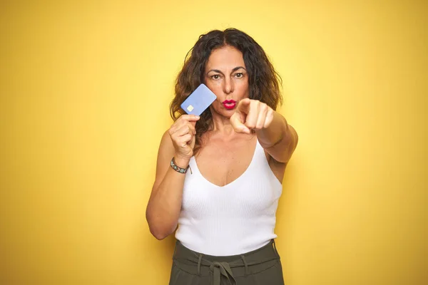 Middle age senior woman holding credit card over yellow isolated background pointing with finger to the camera and to you, hand sign, positive and confident gesture from the front