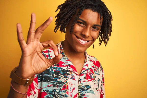 Afro man with dreadlocks on vacation wearing floral shirt over isolated yellow background doing ok sign with fingers, excellent symbol