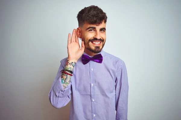 Young man with tattoo wearing purple shirt and bow tie over isolated white background smiling with hand over ear listening an hearing to rumor or gossip. Deafness concept.