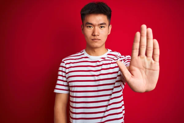 年轻的亚裔男子穿着条纹T恤 站在孤立的红色背景上 停止手心唱着歌 脸上带有消极而严肃的手势的警告表情 — 图库照片