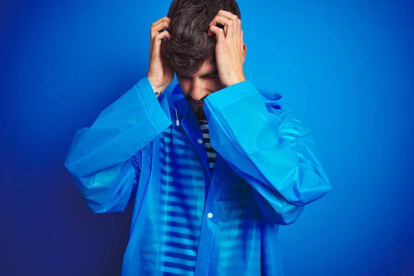Young handsome man wearing rain coat standing over isolated blue background suffering from headache desperate and stressed because pain and migraine. Hands on head.