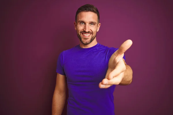 年轻男子穿着宽松的紫色T恤 背负着淡紫色的背景 微笑友好地握手表示问候和欢迎 成功的业务 — 图库照片