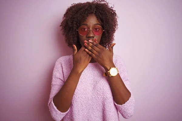孤立したピンクの背景の上にセーターとサングラスを身に着けている若いアフリカのアフロ女性は間違いのための手で口を覆うショックを受けた 秘密概念 — ストック写真