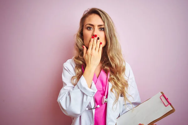 Young beautiful doctor woman holding clipboard over pink isolated background cover mouth with hand shocked with shame for mistake, expression of fear, scared in silence, secret concept