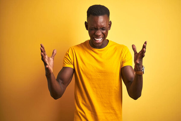 Young african american man wearing casual t-shirt standing over isolated yellow background celebrating mad and crazy for success with arms raised and closed eyes screaming excited. Winner concept