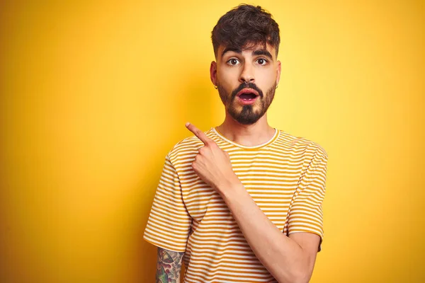 Young man with tattoo wearing striped t-shirt standing over isolated yellow background Surprised pointing with finger to the side, open mouth amazed expression.