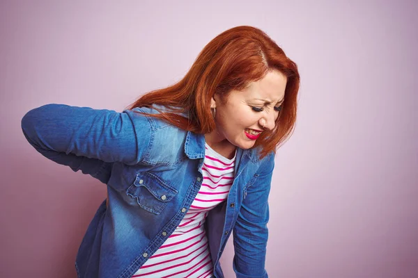 Beautiful redhead woman wearing denim shirt and striped t-shirt over isolated pink background Suffering of backache, touching back with hand, muscular pain