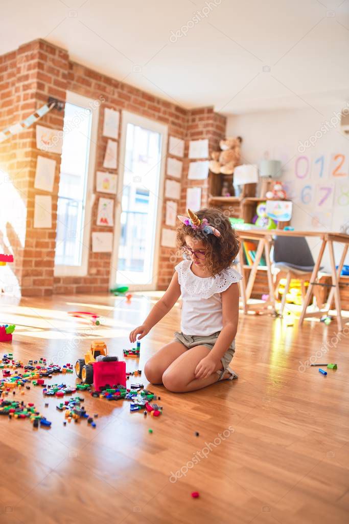 Beautiful toddler wearing glasses and unicorn diadem playing with tractor and building blocks at kindergarten