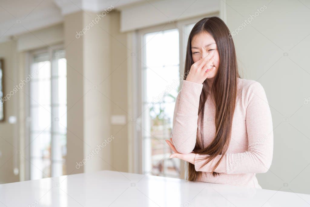 Beautiful Asian woman wearing casual sweater on white table smelling something stinky and disgusting, intolerable smell, holding breath with fingers on nose. Bad smells concept.