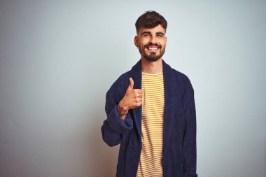 Dövmeli, bornozlu genç bir adam izole edilmiş beyaz arka planda durup mutlu bir el hareketi yapıyor. Kameraya bakıp başarıyı gösteren ifade onaylanıyor.