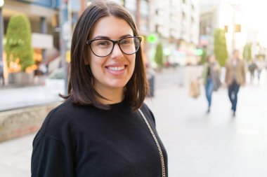 Şehir sokaklarında yürürken heyecanlı gülümseyen güzel genç esmer kadın, şehirde açık havada mutlu ve güven ifade ayakta