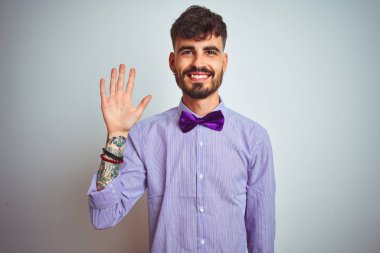Mor gömlekli ve papyonlu genç adam izole edilmiş beyaz arka planda beş numaralı parmaklarıyla işaret ederken kendinden emin ve mutlu gülümsüyor..