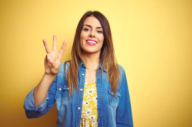 Sarı arka planda duran genç ve güzel bir kadın kendini güvende ve mutlu bir şekilde gülümserken üç numarayı gösteriyor ve işaret ediyor..