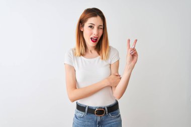 Güzel kızıl saçlı kadın rahat t-shirt izole beyaz arka plan üzerinde duran mutlu yüz zafer işareti yapıyor kameraya göz kırparak gülümseyerek giyiyor. İki numara.