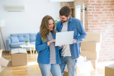 Yeni bir daireye taşımak için mutlu karton kutular etrafında bir odada bilgisayar laptop ayakta kullanarak genç çift