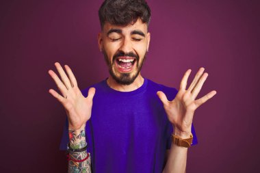 İzole edilmiş mor arka plan üzerinde duran dövmeli genç adam kolları havada ve gözleri kapalı heyecanla haykırarak başarının çılgınlığını kutluyor. Kazanan konsept