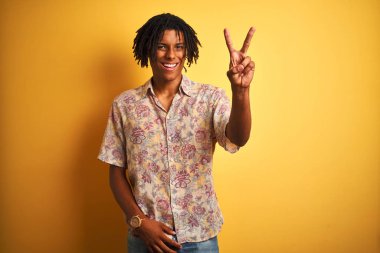 Tatilde saçı rastalı bir Afro adam çiçekli bir gömlek giyer ve izole edilmiş sarı bir arkaplanda güler yüzlü bir şekilde kameraya bakıp zafer işareti yapar. İki numara..