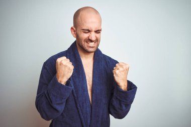 Mavi bornoz giyen, rahat bir yaşam tarzı olan, izole edilmiş arka planda çok mutlu ve heyecanlı bir şekilde kollarını kaldırıp, gülümseyerek ve başarı için çığlık atarak kazanan bir jest yapan genç adam. Kutlama konsepti.