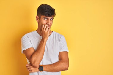 İzole edilmiş sarı arka plan üzerinde duran beyaz tişörtlü genç Hintli adam elleri ağız yiyen tırnaklarla gergin ve stresli görünüyor. Endişe sorunu.