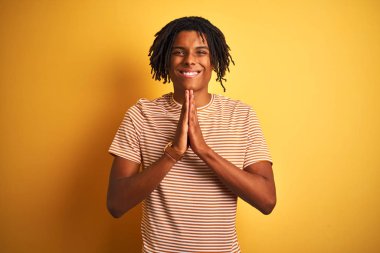 Sarı arka planda duran çizgili tişörtlü Afro bir adam ellerini birleştirip af diliyor kendinden emin bir şekilde gülümsüyor..