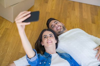 Yeni evin zemininde yatan genç çift akıllı telefon kullanarak selfie fotoğrafı çekerek, yeni daireye taşındığı için mutlu bir şekilde gülümsüyor