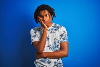 Rasta desenli tişörtlü Afro-Amerikan bir adam izole edilmiş mavi arka plan üzerine çiçekli tişört giyiyor ve kolları çaprazlanmış depresyondan sıkılmış ve yorgun görünüyor..
