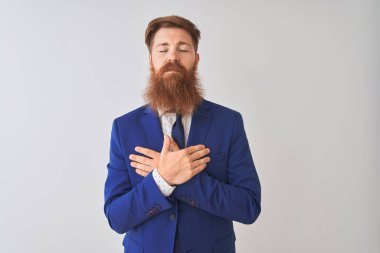 İzole edilmiş beyaz arka planda duran takım elbiseli genç kızıl saçlı işadamı elleri göğsünde, gözleri kapalı ve yüzünde minnet dolu bir jest ile gülümsüyor. Sağlık kavramı.