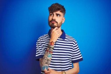 Çizgili polo giyen genç bir adam soyutlanmış mavi arka planda duruyor ve elleri çenesinde soru sormayı düşünüyor, düşünceli bir ifade. Düşünceli bir yüzle gülümsüyor. Şüphe kavramı.